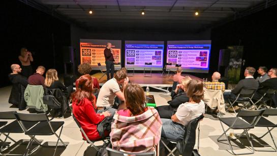 People are gathered in a workshop setting. Someone is presenting on the stage. 