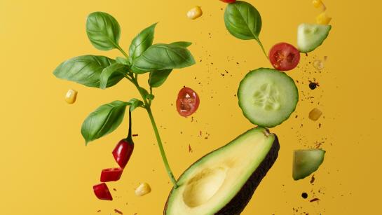 Floating healthy food including cucumber, avocado, basil and red pepper against a yellow background