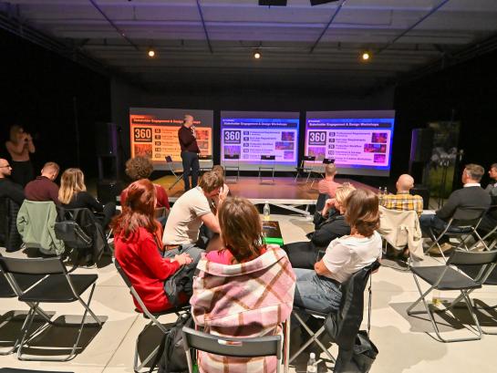 People are gathered in a workshop setting. Someone is presenting on the stage. 