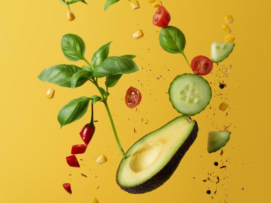 Floating healthy food including cucumber, avocado, basil and red pepper against a yellow background