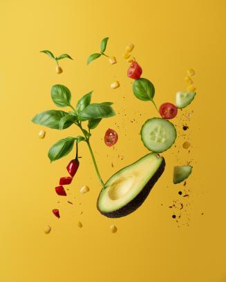 Floating healthy food including cucumber, avocado, basil and red pepper against a yellow background